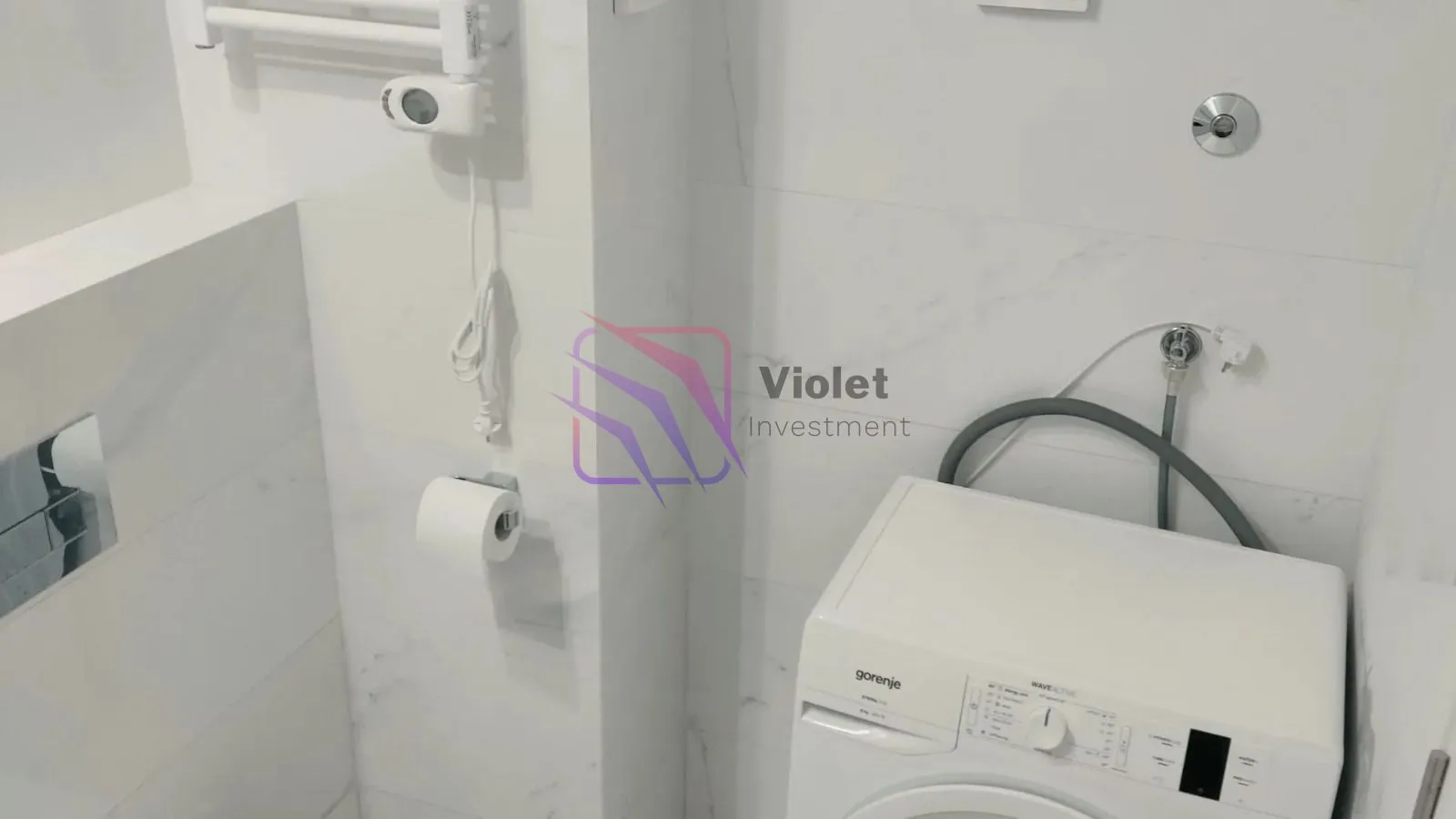 Kupatilo sa bijelim pločicama, toaletnim papirom i veš mašinom.