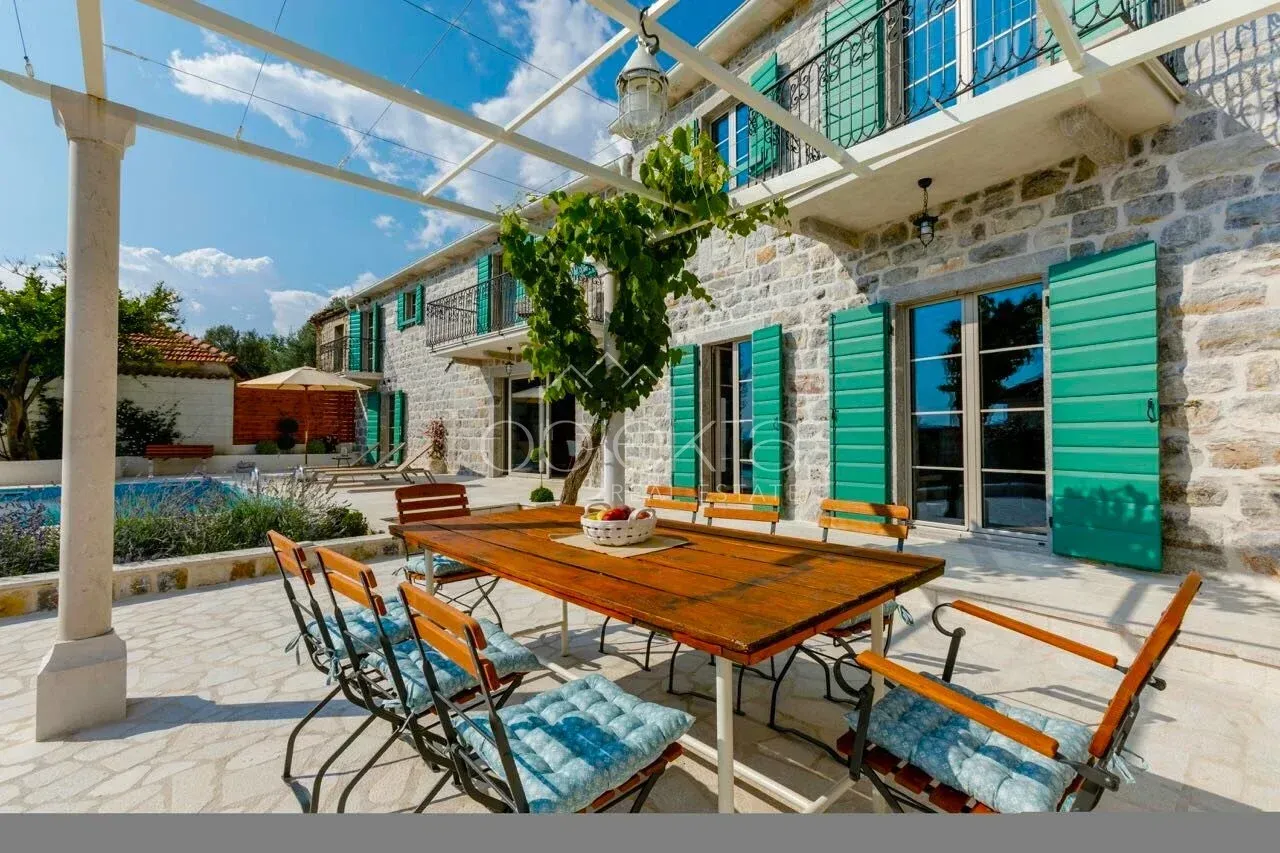 Terrace with a wooden table and chairs in front of a stone house with a swimming pool.
