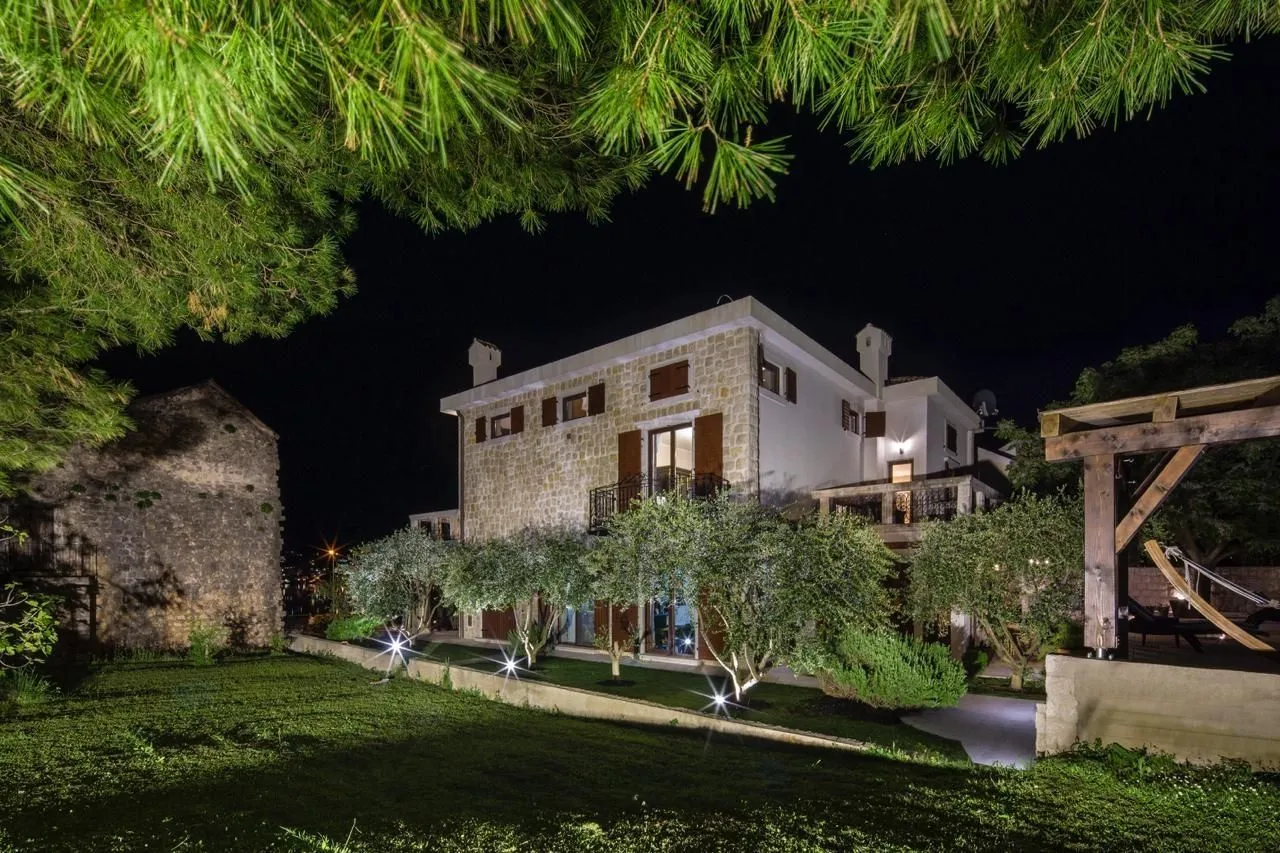 A stone house with an illuminated yard and olive trees at night.