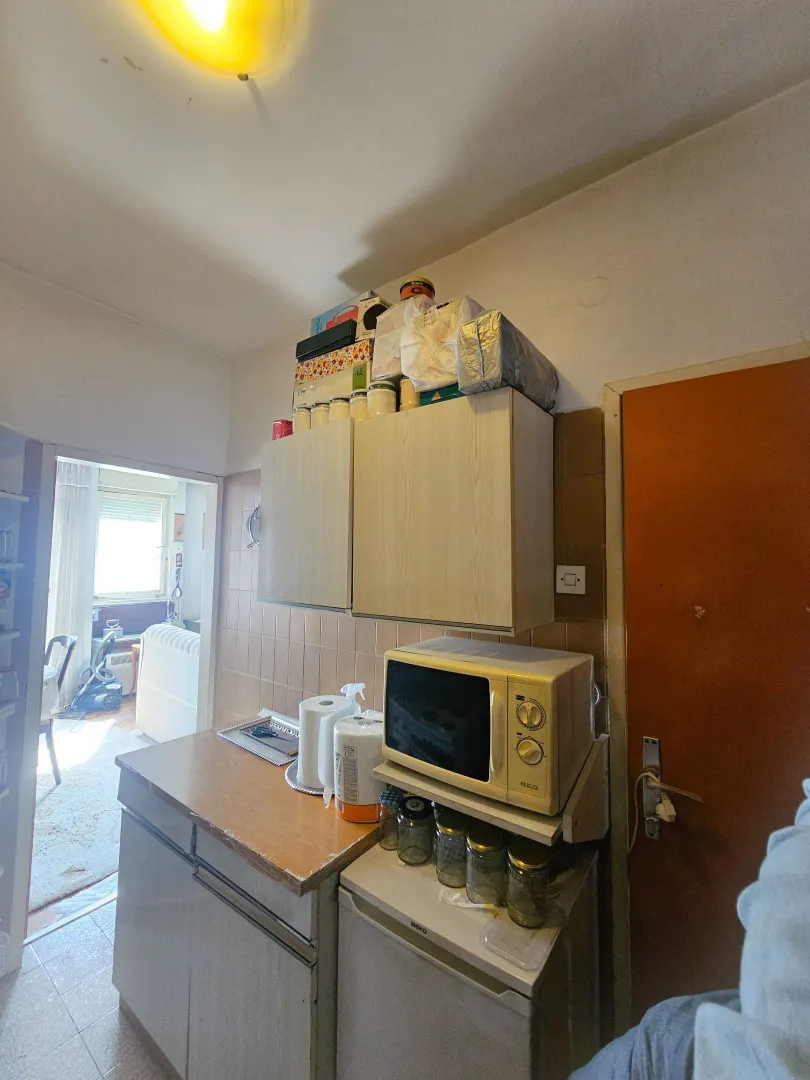 Starija i neuredna kuhinja sa ormarićima, mikrotalasnom pećnicom i otvorenim vratima prema drugoj prostoriji.