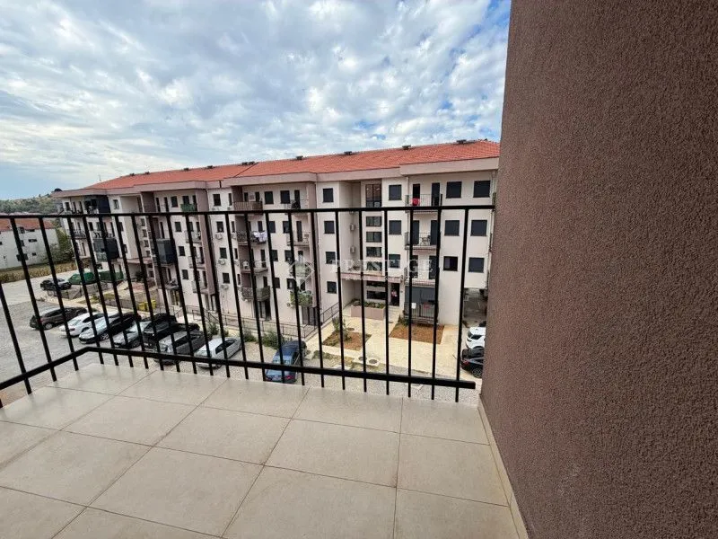 Pogled sa balkona na zgradu i parking sa parkiranim automobilima.