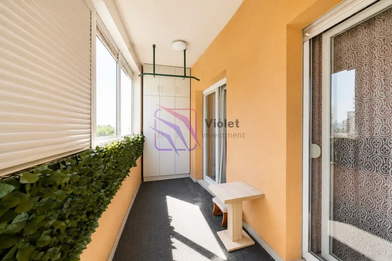Zastakljeni balkon sa malim stolom, ormarom i vještačkom travom na ogradi.
