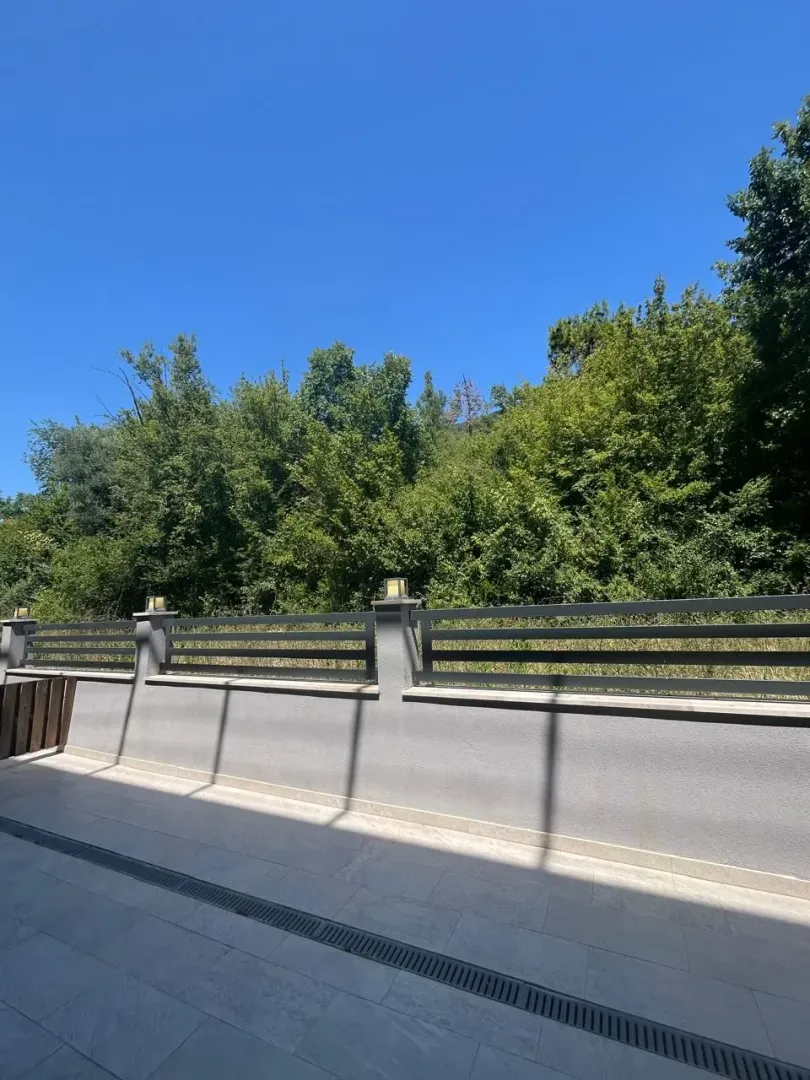 Popločano dvorište sa ogradom i pogledom na zelenilo.