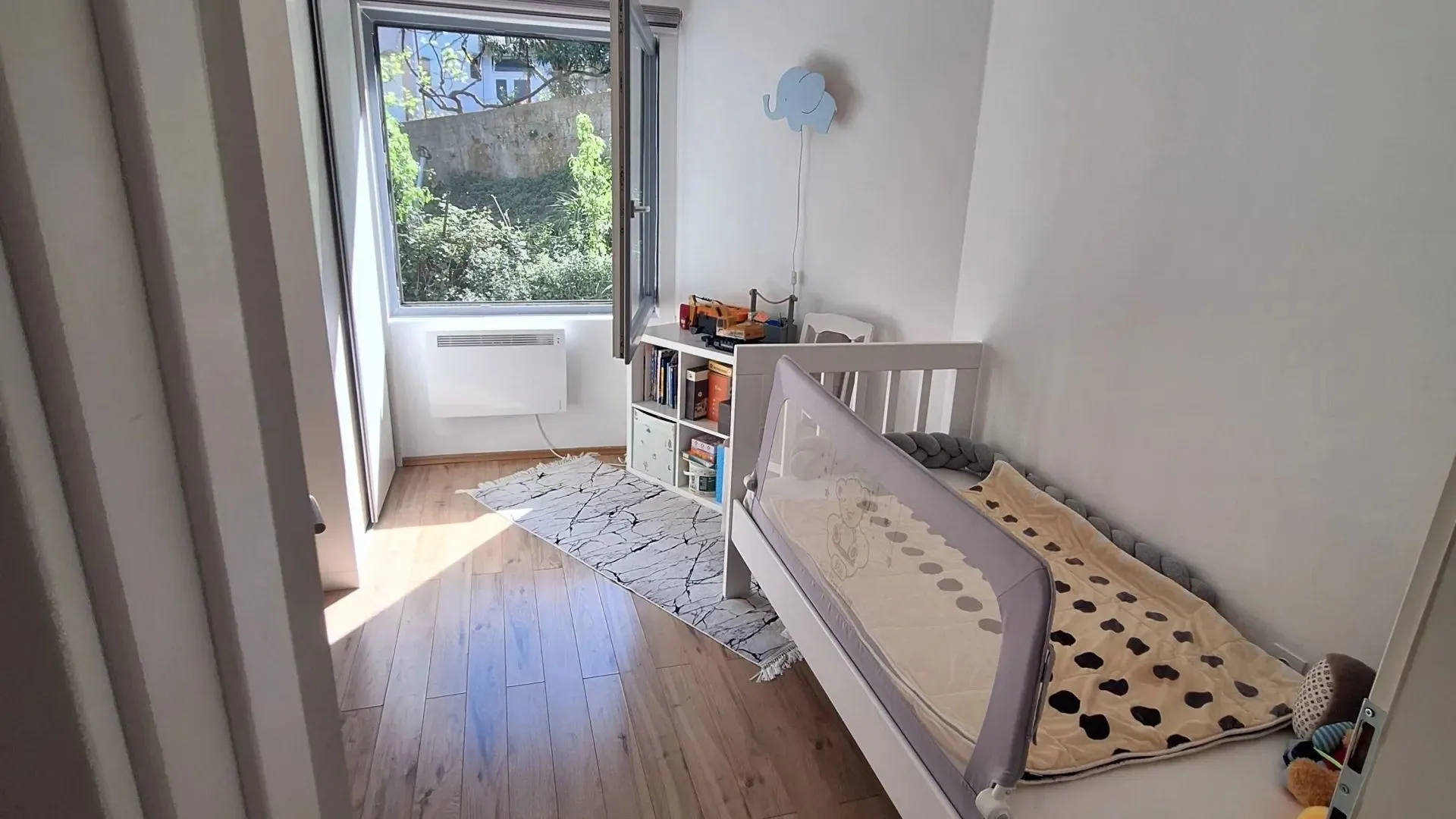 Children's bedroom with a bed, bookcase, and a window with a green view.
