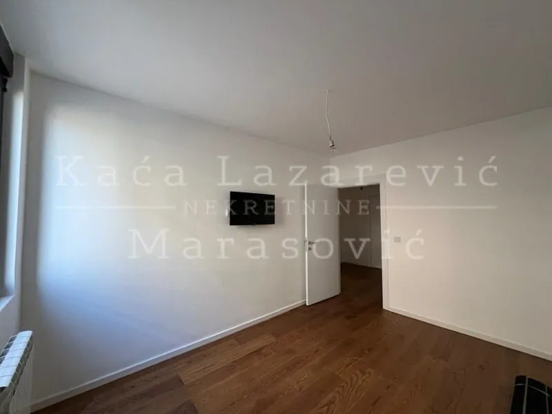 Prazna soba sa belim zidovima, drvenim podom i televizorom na zidu.