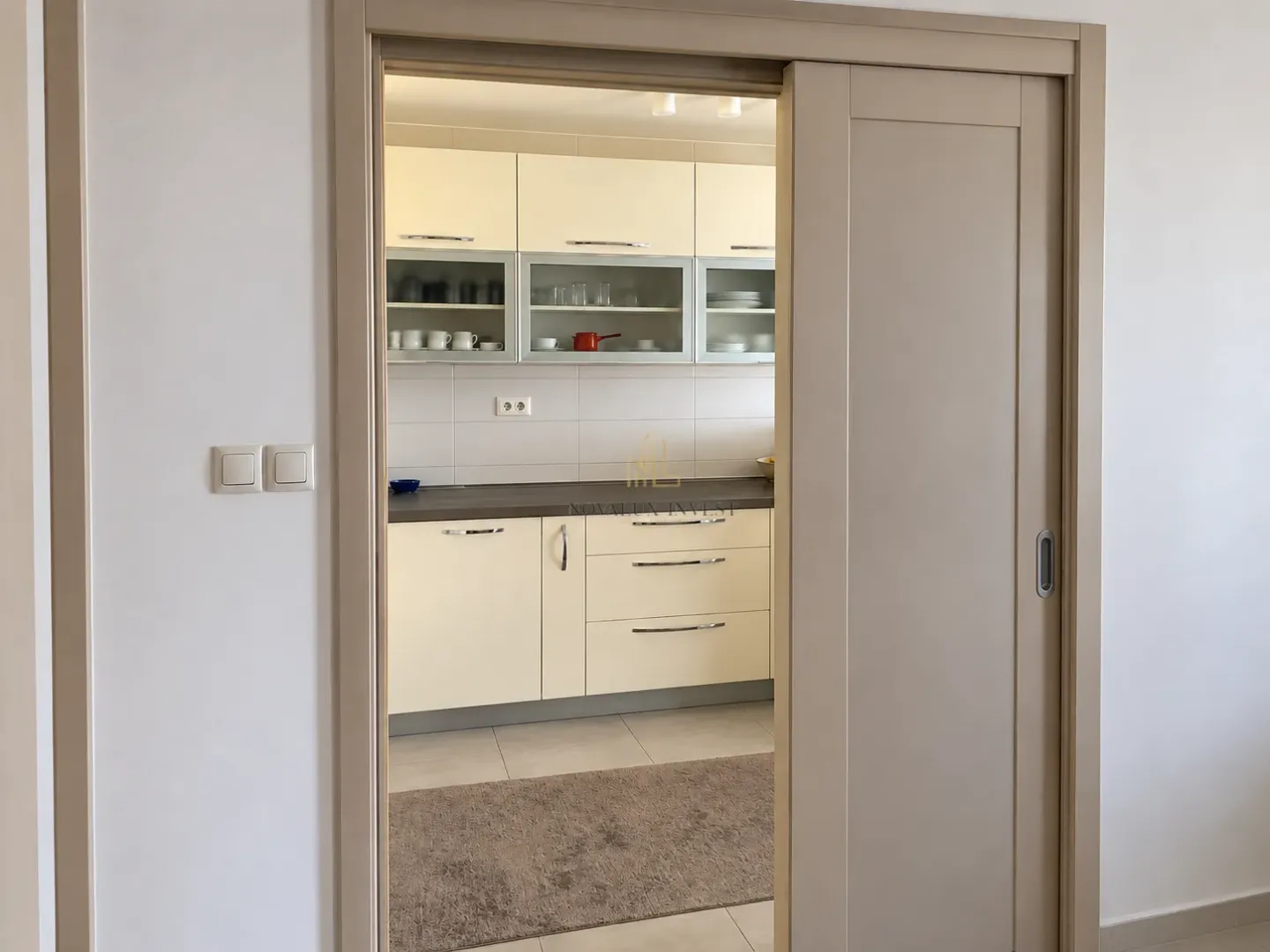 View through a sliding door into a fitted kitchen with beige cabinets.