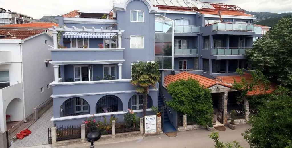 Prednji pogled na višespratnu plavu zgradu sa terasama i crvenim krovom.