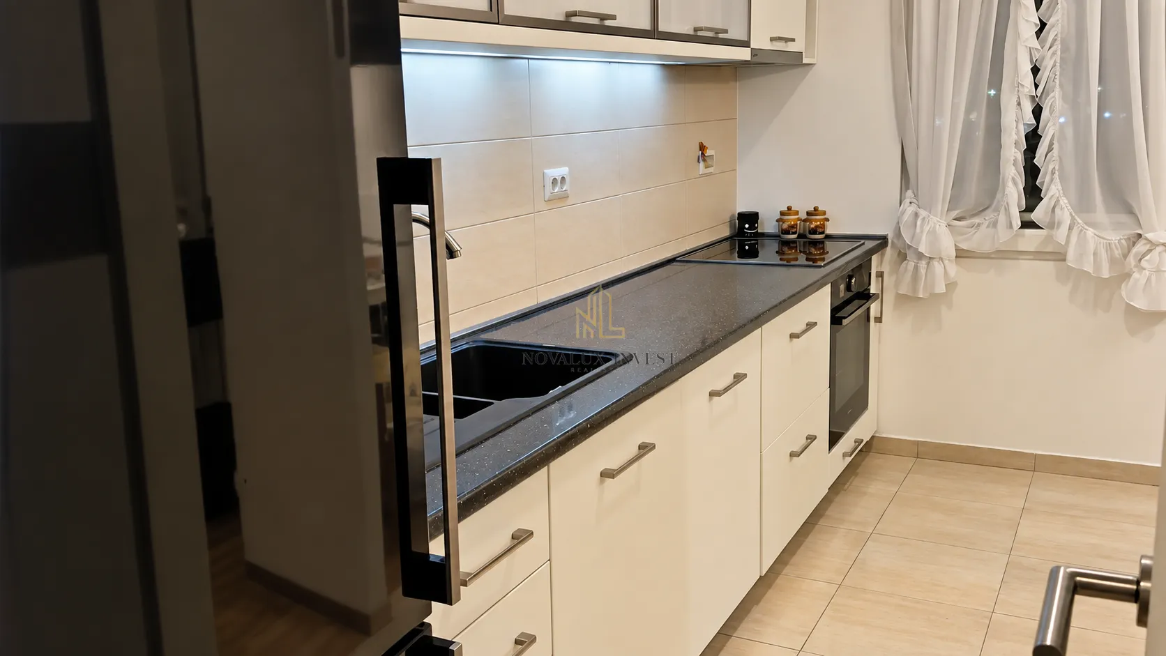 Pogled na moderno opremljenu kuhinju sa bijelim elementima i tamnom radnom površinom.