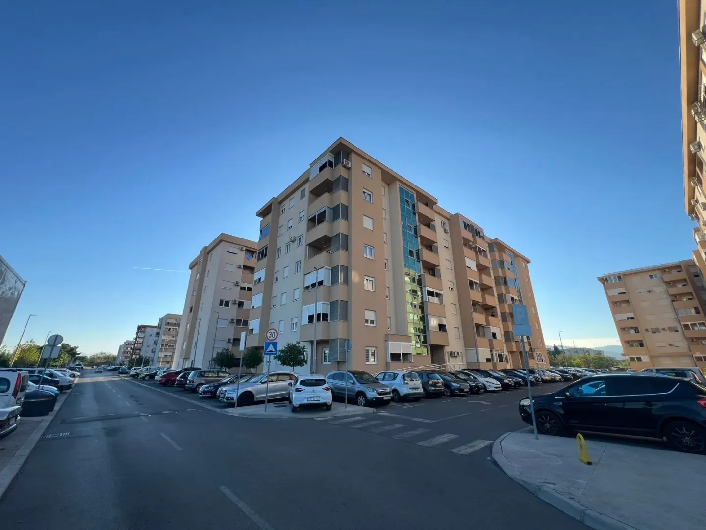 Pogled na višestambenu zgradu i okolni parking ispred ulaza.