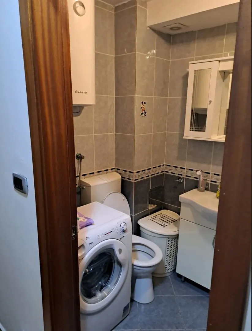 Manje kupatilo sa sanitarijama, veš mašinom i bijelim ormarićem sa ogledalom.