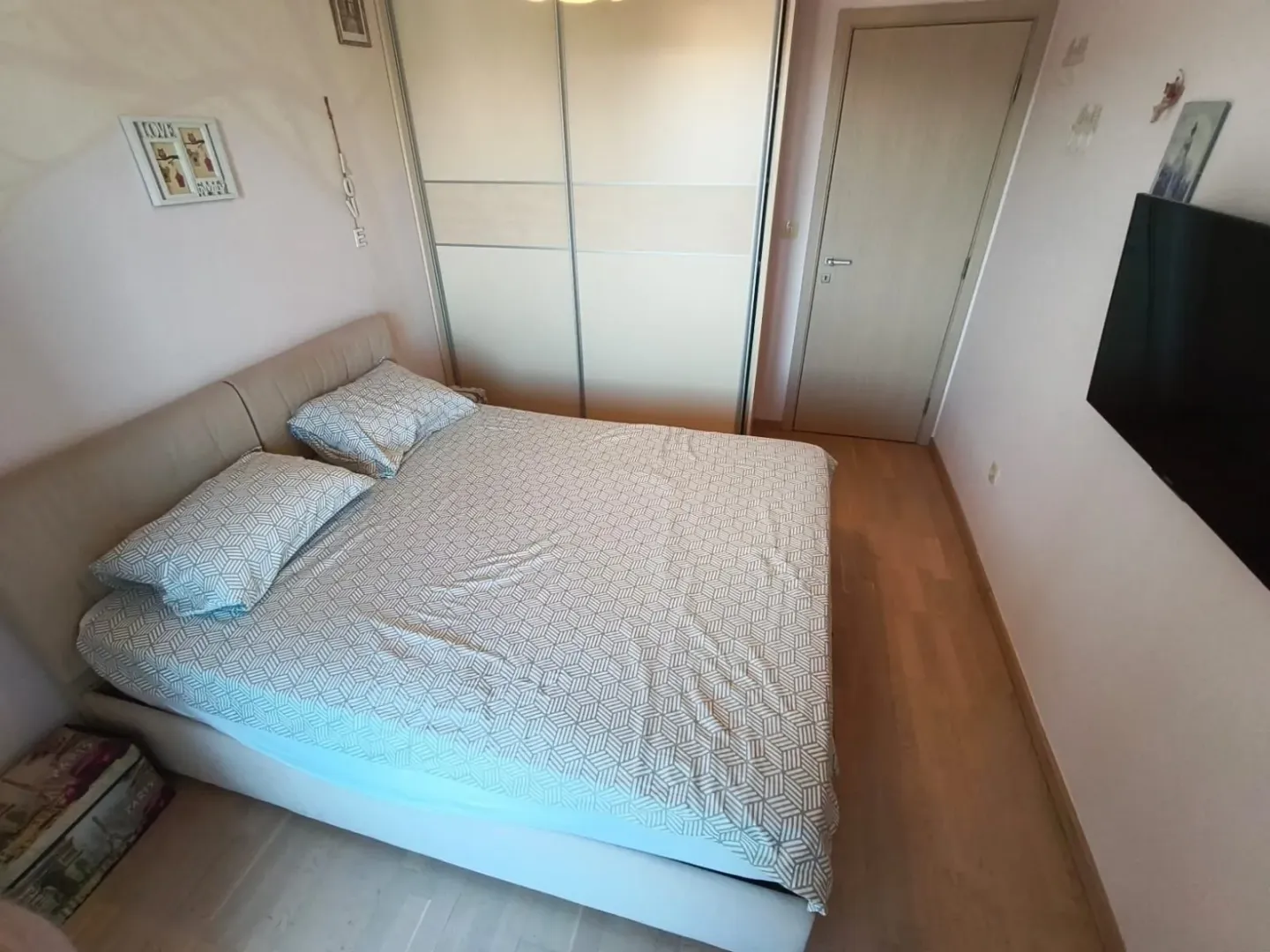 Spavaća soba sa bračnim krevetom, plakarom i televizorom na zidu.