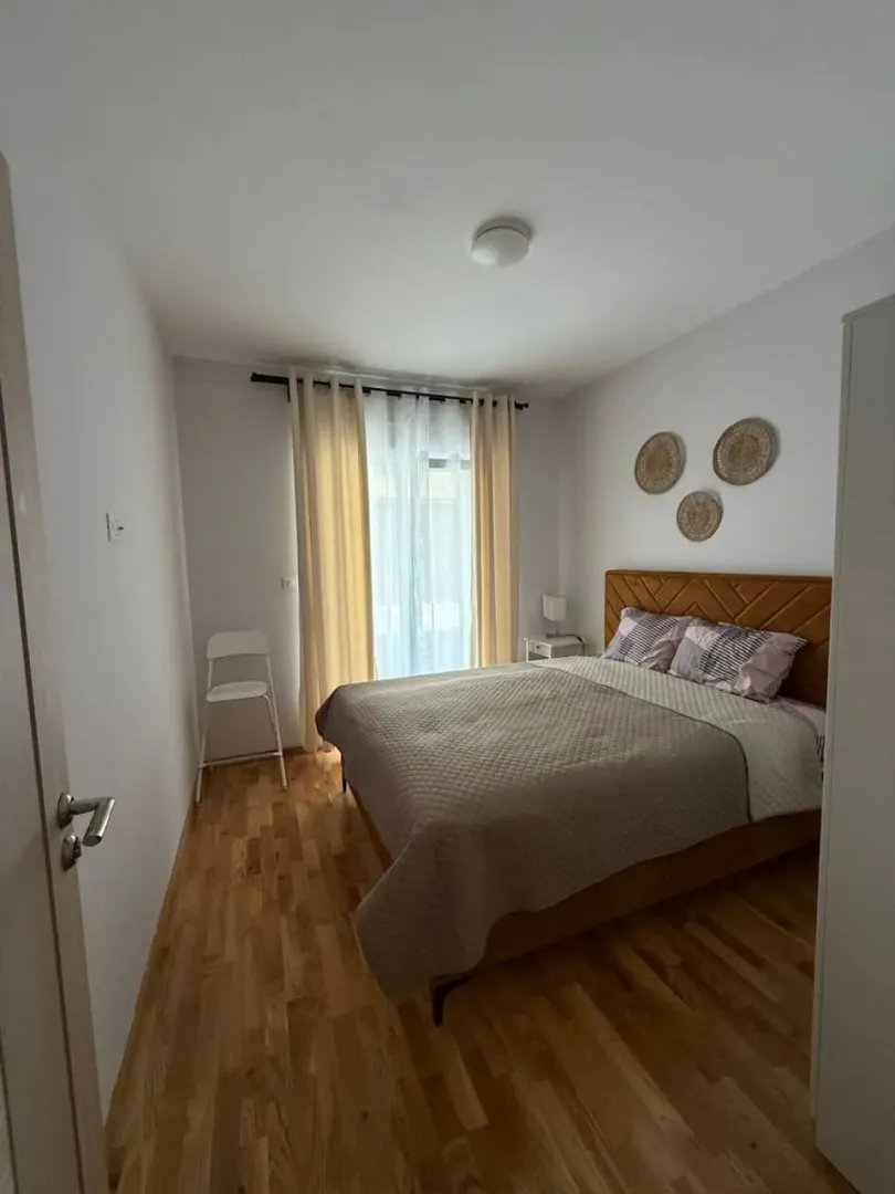 Uređena spavaća soba sa bračnim krevetom, drvenim podom i prozorom sa zavjesama.