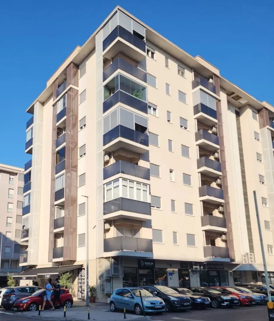 Pogled na spoljašnost stambene zgrade sa terasama i parkiranim automobilima ispred.