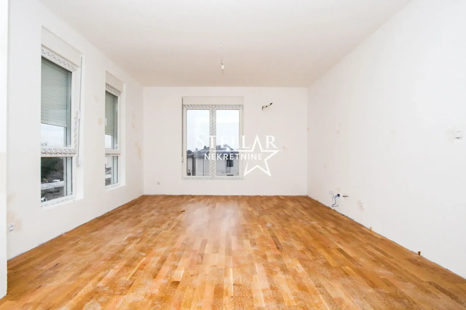 Empty room with white walls, wooden parquet flooring, and windows.