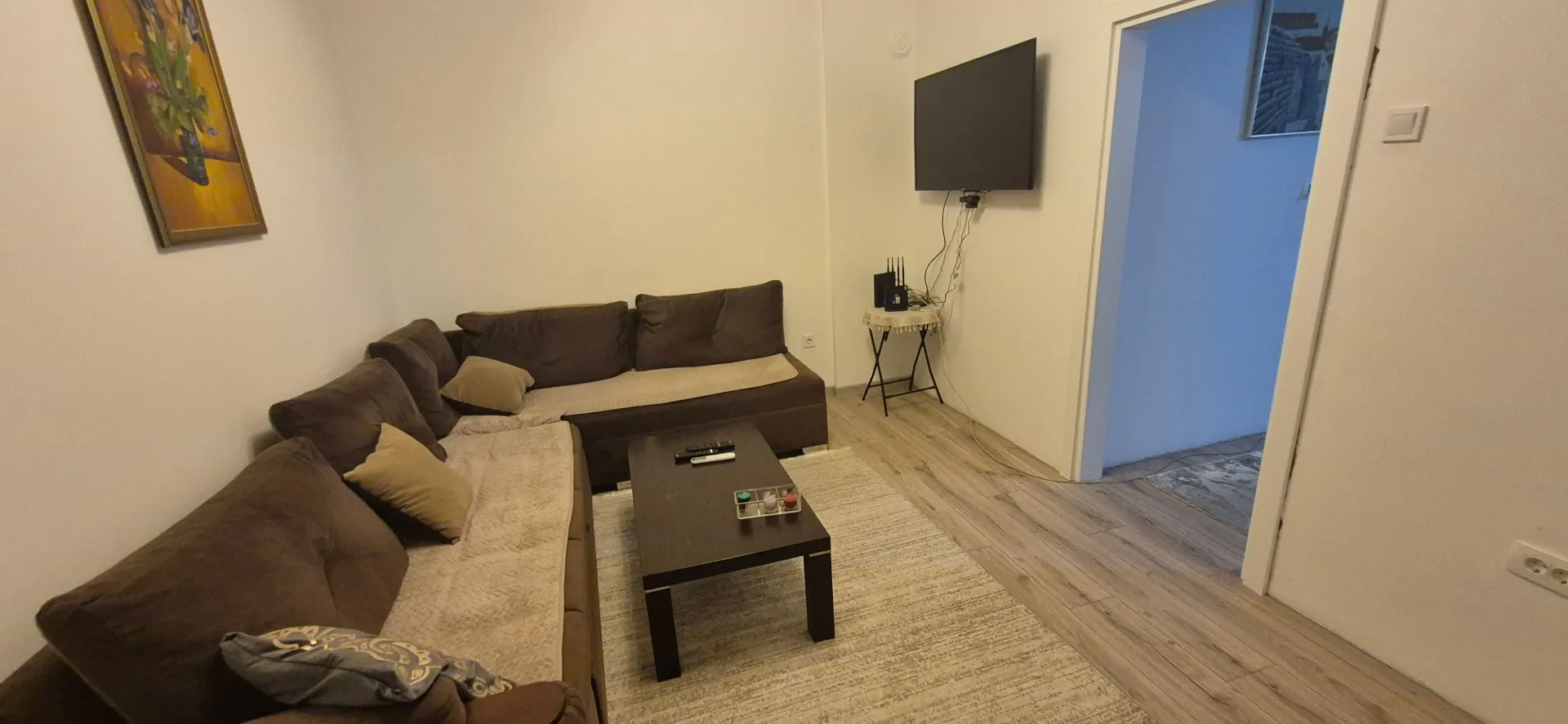 Dnevna soba sa ugaonom garniturom, stočićem i televizorom na zidu.