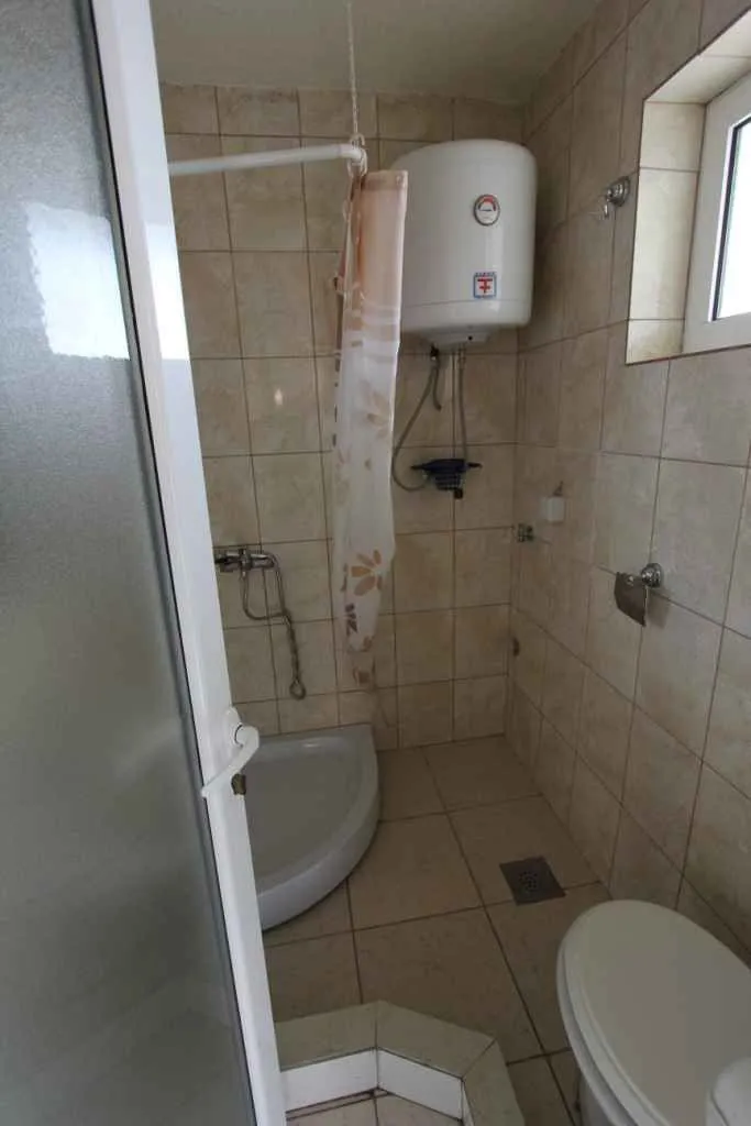 Manje kupatilo sa tuš kabinom, bojlerom i WC šoljom obloženo keramičkim pločicama.