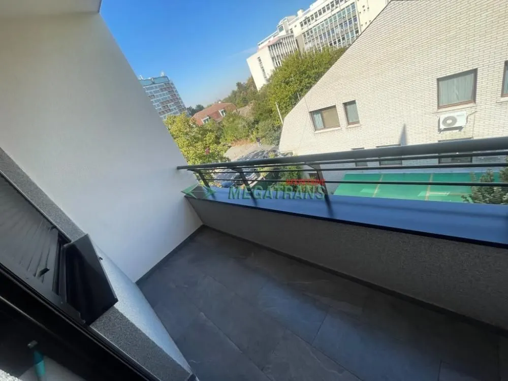 Manji popločani balkon sa metalnom ogradom i pogledom na okolne zgrade i zelenilo.