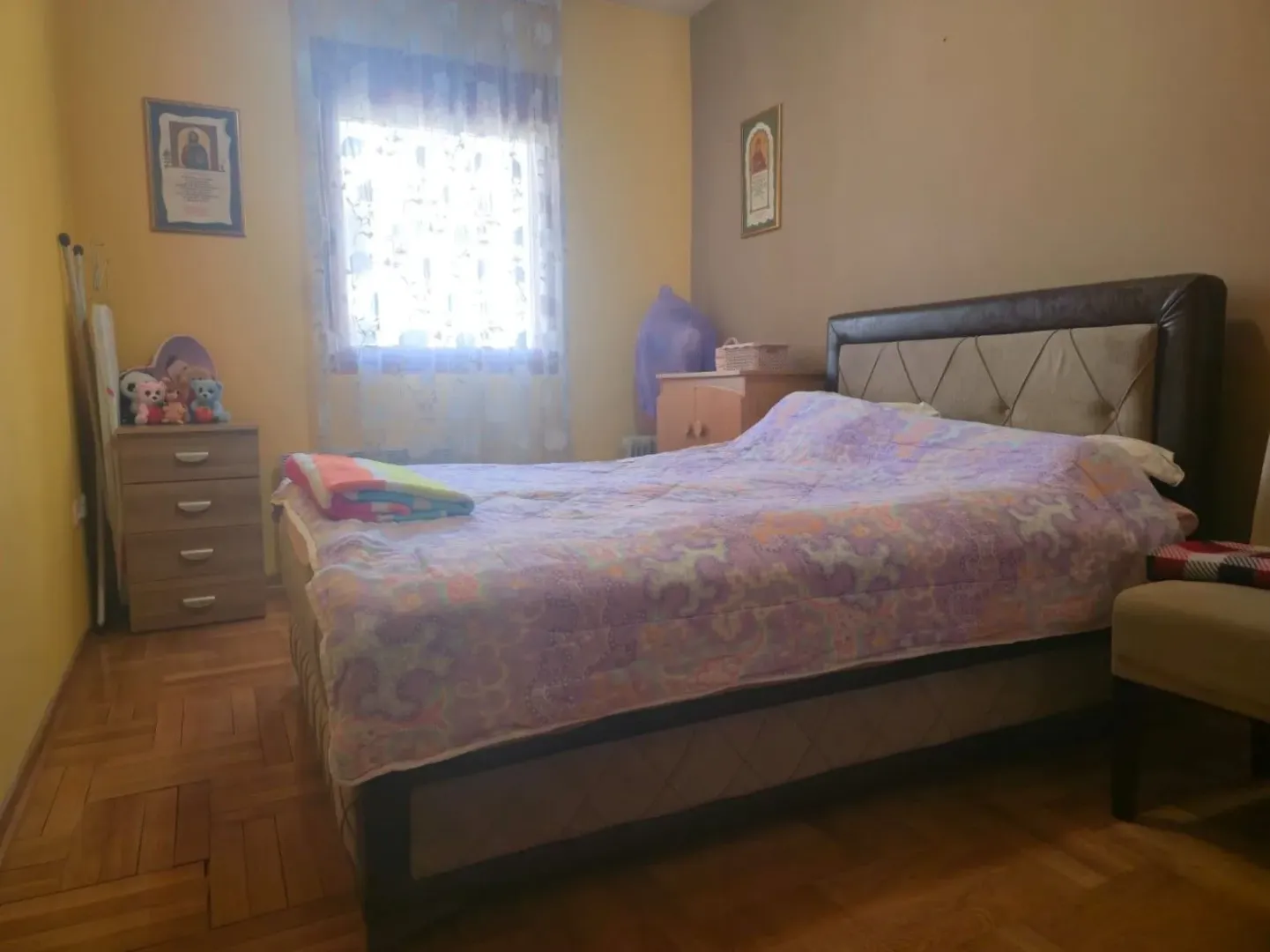 Namještena spavaća soba sa bračnim krevetom, komodom i prozorom.