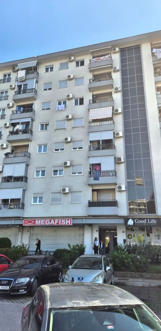 Spoljašnji izgled stambene zgrade sa poslovnim prostorima u prizemlju i parkiranim automobilima ispred.