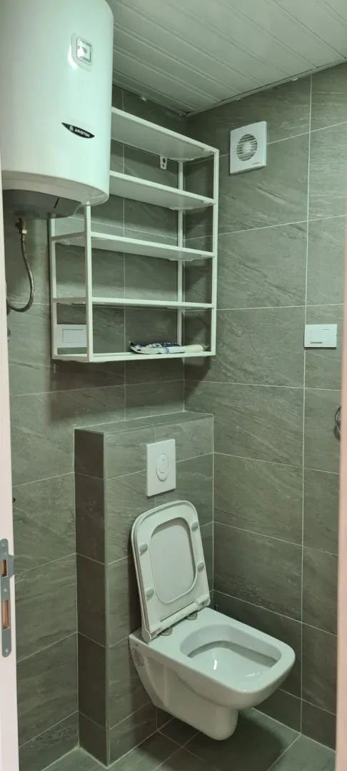 Mali toalet sa sivim pločicama, visećom šoljom i bojlerom.
