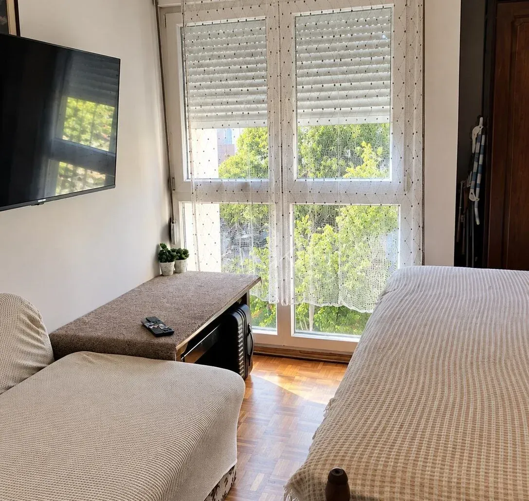 Manja spavaća soba sa krevetom, foteljom, radnim stolom i televizorom na zidu.