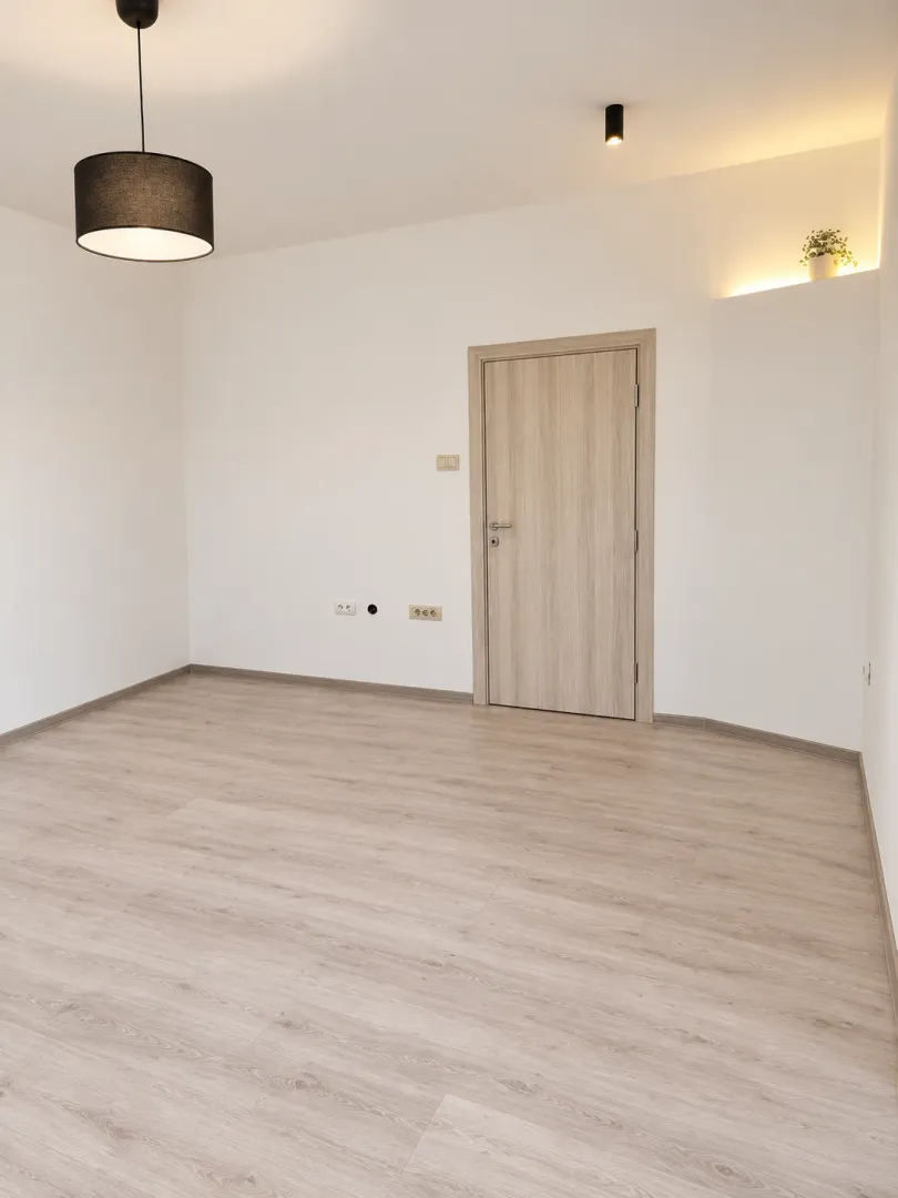 An empty room with light laminate flooring, white walls, and a wooden door.