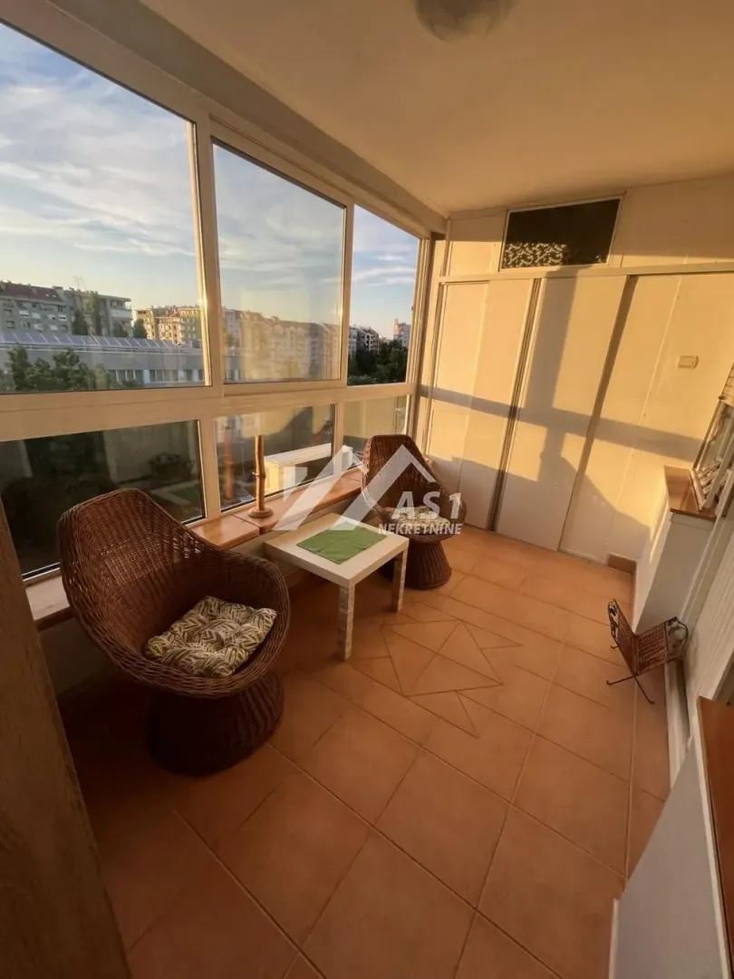 Zastakljena terasa sa pletenim foteljama i pogledom na grad.