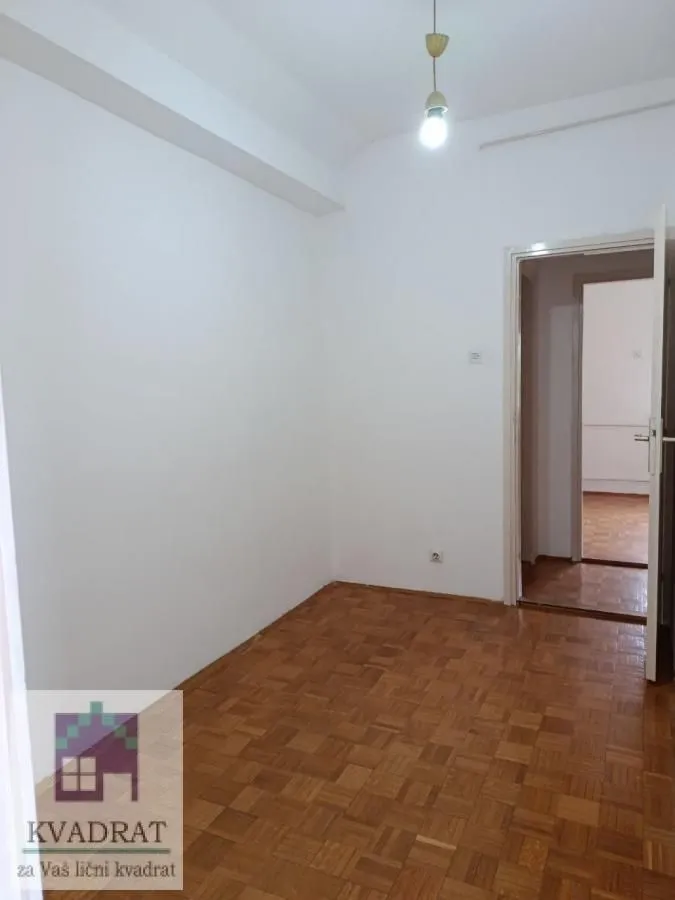Prazna soba sa belim zidovima, parketom i otvorenim vratima.