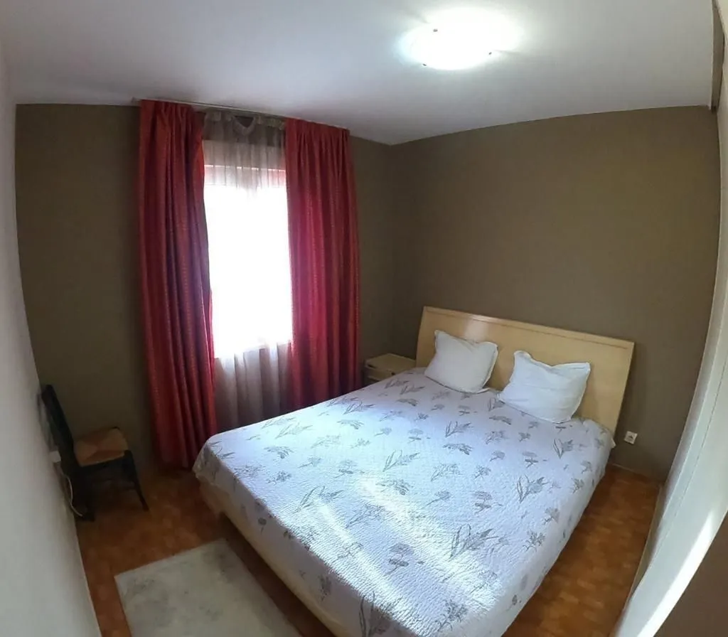 Jednostavno namještena spavaća soba sa bračnim krevetom i crvenim zavjesama.