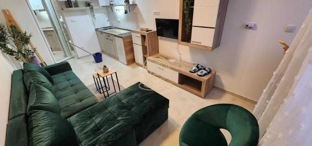 Dnevna soba sa garniturom, televizorom i kuhinjom u pozadini.