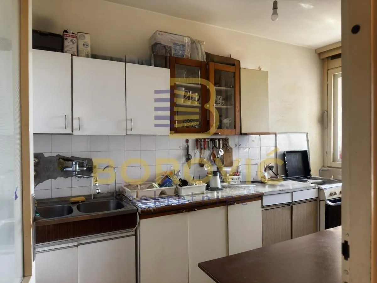 Starija kuhinja sa belim i drvenim elementima, sudoperom i šporetom.
