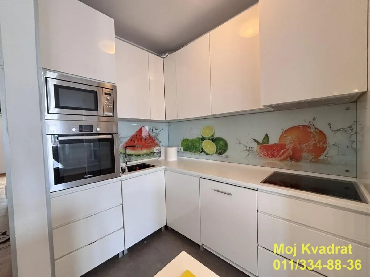 Moderna bela kuhinja sa ugrađenom rernom, mikrotalasnom i dekorativnom pozadinom sa motivom voća.