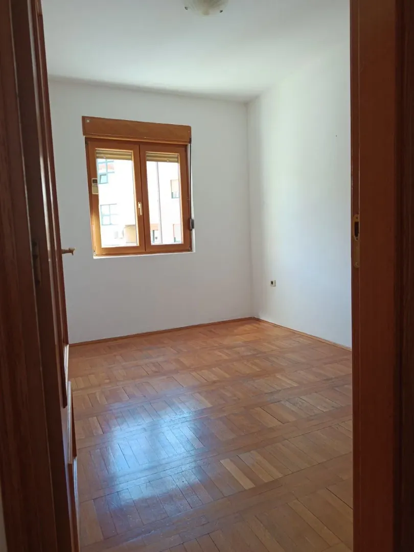 Empty room with wooden parquet flooring and a window, viewed through an open doorway.