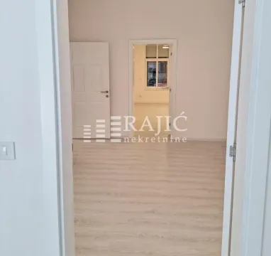 Prazna svetla prostorija sa svetlim laminatom i otvorenim vratima prema drugoj sobi. - image 5