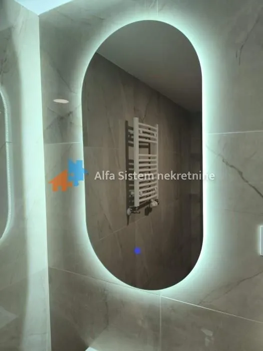 Close-up of the bathroom featuring a large oval LED backlit mirror and ceramic wall tiles.