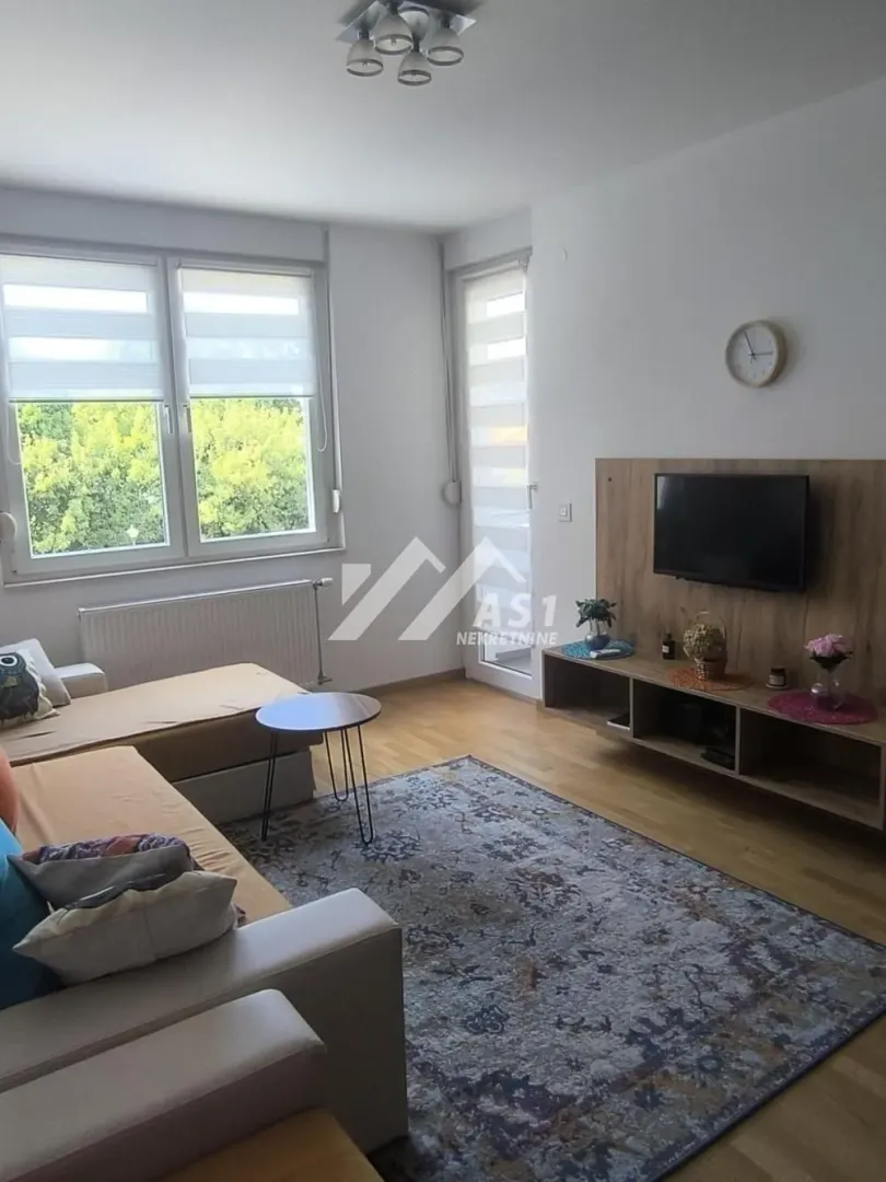 Dnevna soba sa garniturom, TV komodom i velikim prozorima.