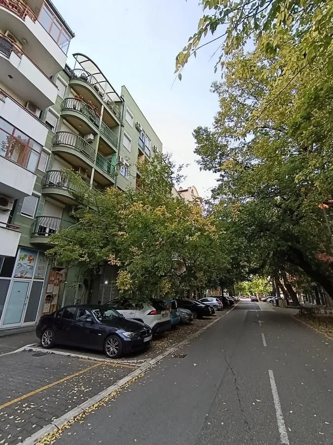 Pogled na ulicu ispred zgrade sa parkiranim automobilima i drvećem.