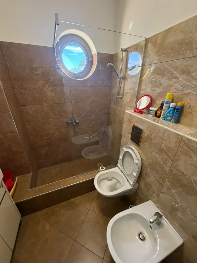 Modern bathroom with a walk-in shower, toilet, bidet, and a round window.