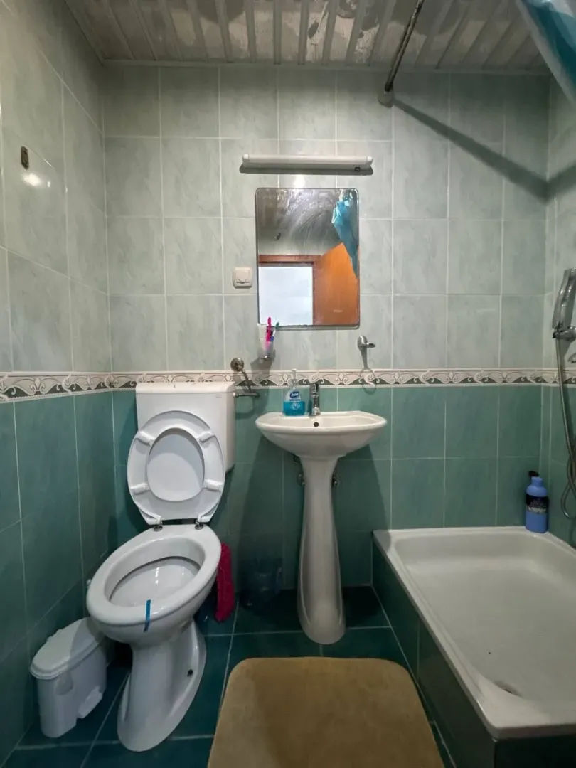 Manje kupatilo sa zelenim pločicama, umivaonikom, toaletom i tuš kadom.