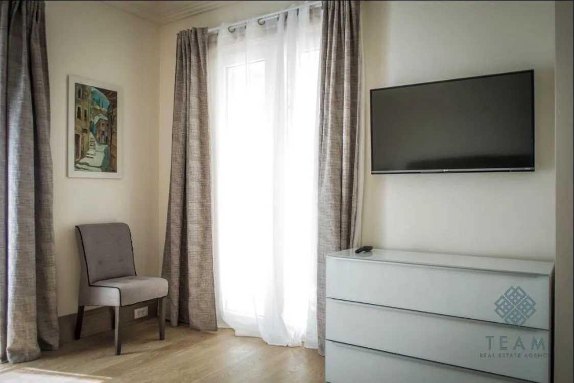 Dnevna soba sa televizorom na zidu, komodom i balkonskim vratima.
