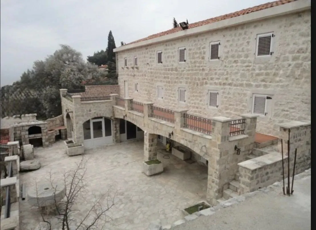 Pogled na kamenu fasadu kuće i popločano dvorište sa terasom i kaminom.