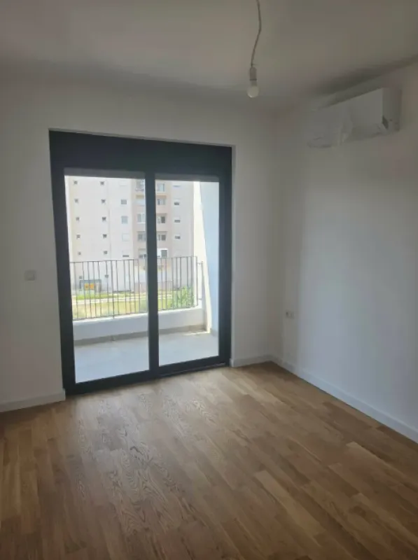 Prazna soba sa drvenim podom, klima-uređajem i izlazom na balkon.