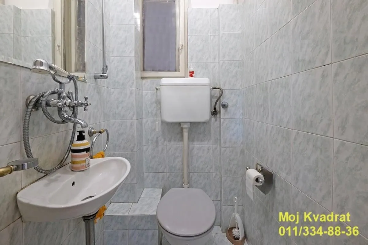 Manje kupatilo obloženo svetlim pločicama sa umivaonikom i wc šoljom.