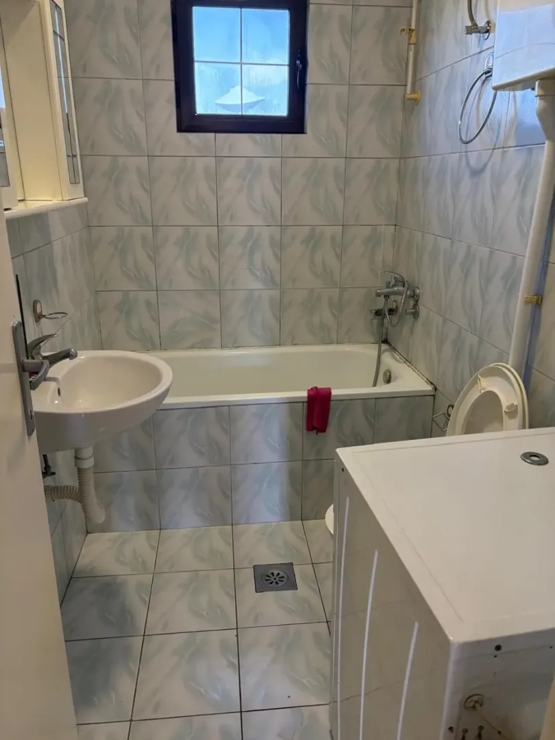 Manje kupatilo obloženo keramičkim pločicama sa kadom, umivaonikom i veš mašinom.