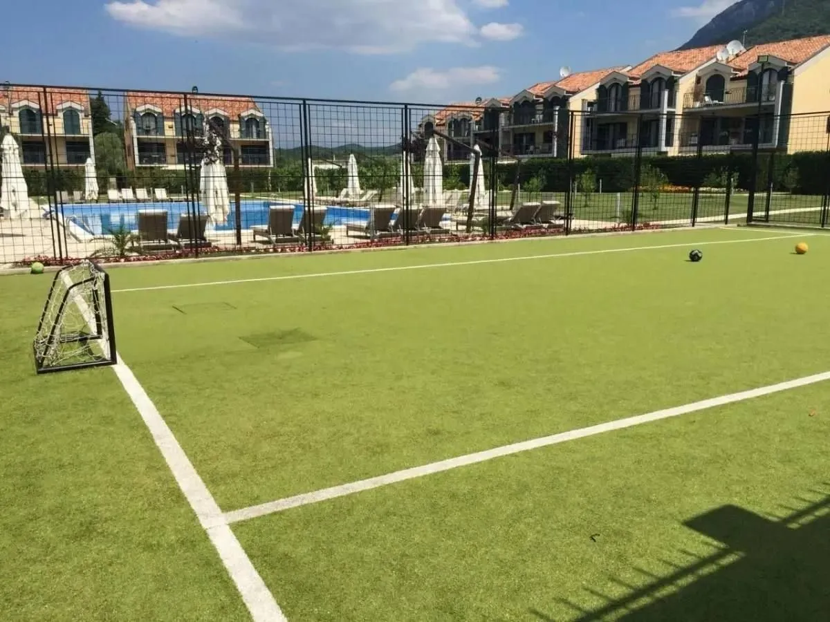 Sportski teren na otvorenom sa vještačkom travom i pogledom na kompleks zgrada sa bazenom.