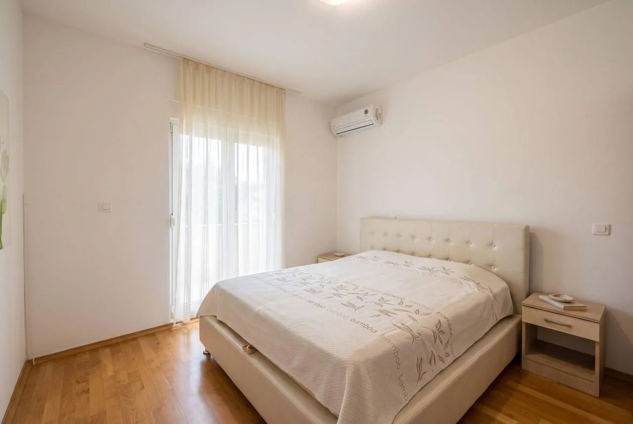 Svjetla spavaća soba sa bračnim krevetom, drvenim podom i izlazom na balkon.