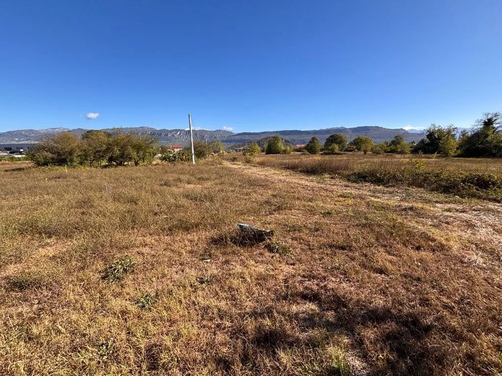 Pogled na ravno građevinsko zemljište sa niskim rastinjem i planinama u pozadini.