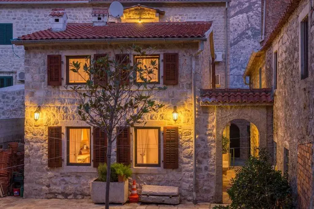 Kamena kuća sa osvijetljenim prozorima i drvenim škurama u večernjim satima.