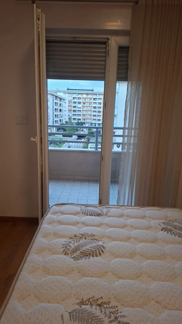 Pogled iz spavaće sobe sa krevetom prema balkonu i zgradama u okruženju.