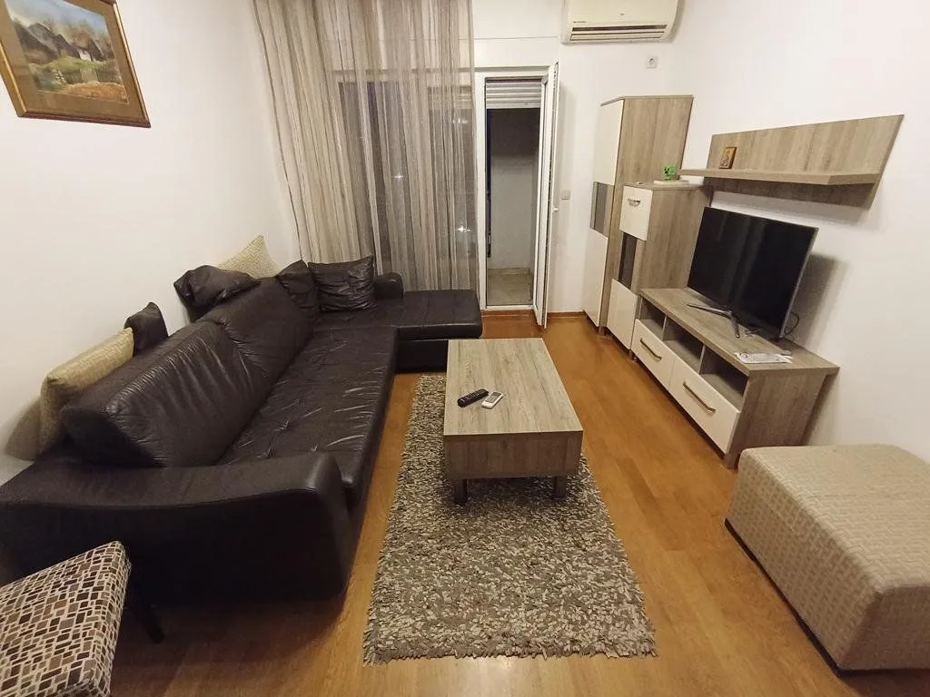 Dnevna soba sa ugaonom garniturom, televizorom i izlazom na balkon.